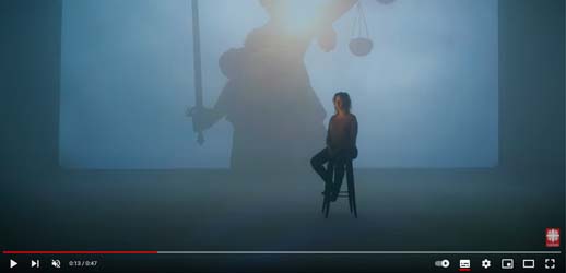 Ausschnitt aus einem Youtube-Video. Frau sitzt auf einem Hocker und hat die Augen geschlossen. Hinter ihr eine Leinwand mit einer Statue, der Justitia. 