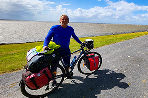 Älterer Mann im blauen Longshirt steht mit seinem gepacktem Fahrrad vor dem Meer und lächelt in die Kamera