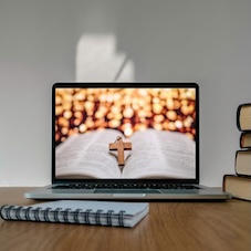 Ein Laptop liegt offen auf einem Schreibtisch. Auf dem Bildschirm ist ein offenes Buch zusehen, auf dem ein braunes Holz liegt.