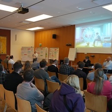 Blick in einen Raum des Hack-a-thons in Kassel. Man sieht die Hinterk&ouml;pfe der Zuschauer sowie einen Referenten vor Flipcharts mit Ergebnissen