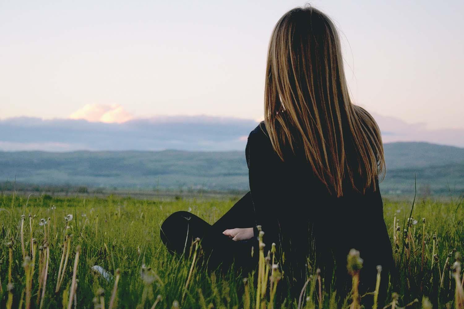 Blick von hinten auf eine langhaarige Frau, die auf einer Wiese im Gras sitzt